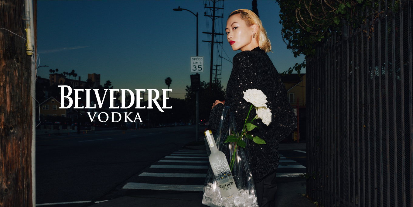 Superchouette- Une personne en tenue noire se tient sur un passage piéton au crépuscule, tenant un sac transparent contenant une bouteille de Vodka Belvedere, de la glace et des fleurs blanches. Un texte en gros caractères indique Belvedere Vodka sur le côté gauche de l'image.